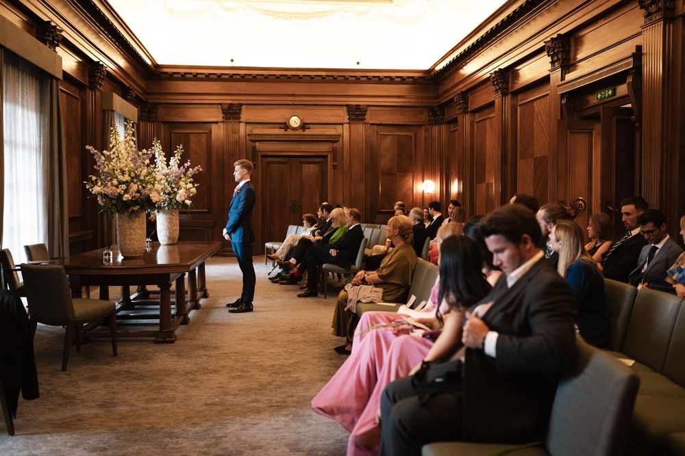 Wedding ceremony guests seated in wood-paneled reception hall with high ceilings.