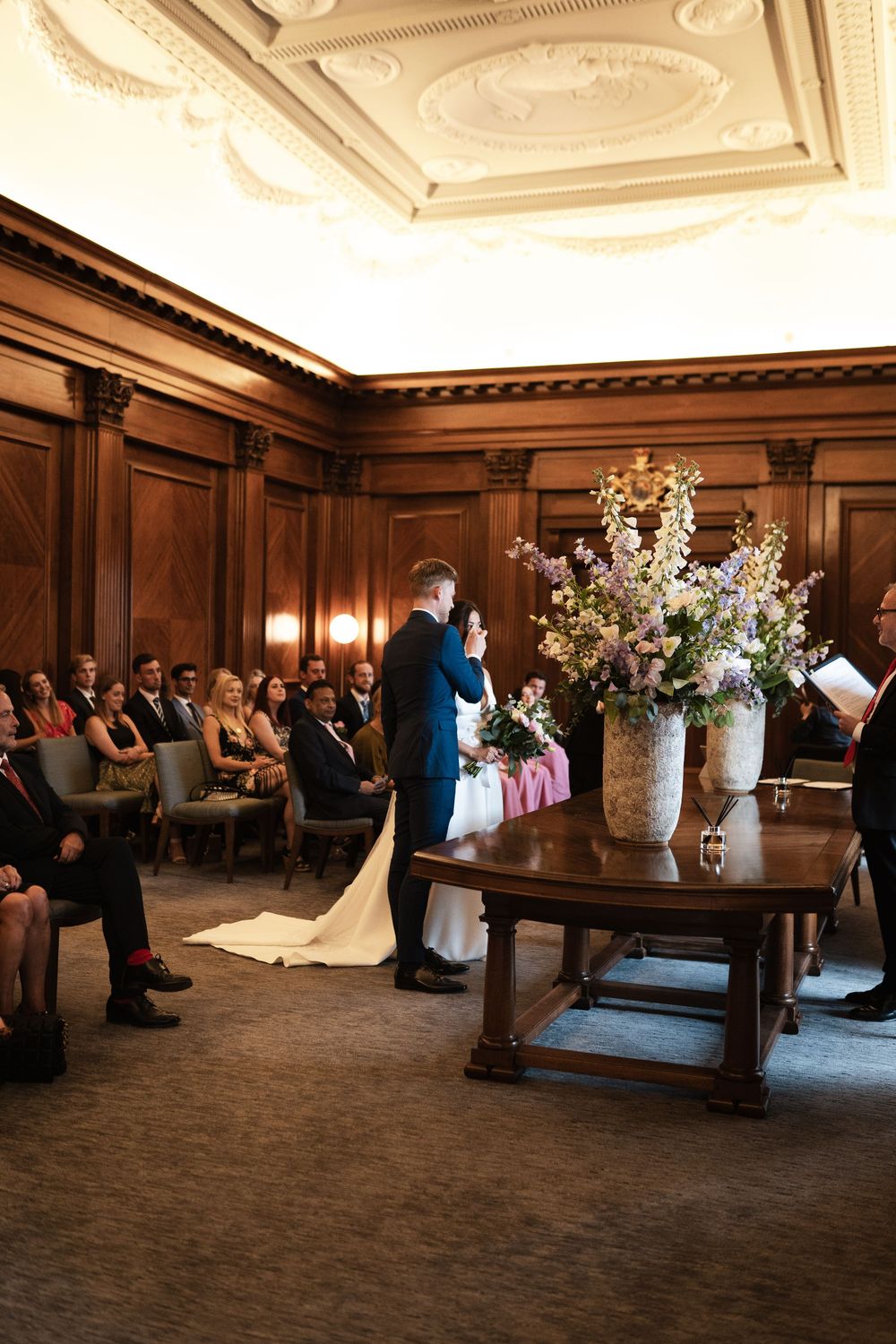 A wedding ceremony in a grand room with wood paneling, ornate ceiling, and stunning floral arrangements.