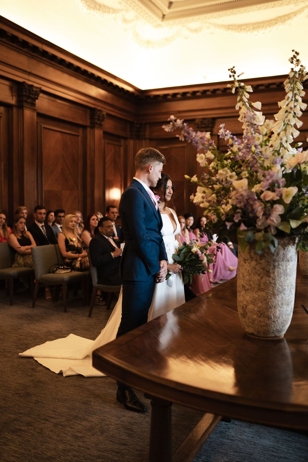 A wedding ceremony takes place in a classic wood-paneled room with elegant floral arrangements.