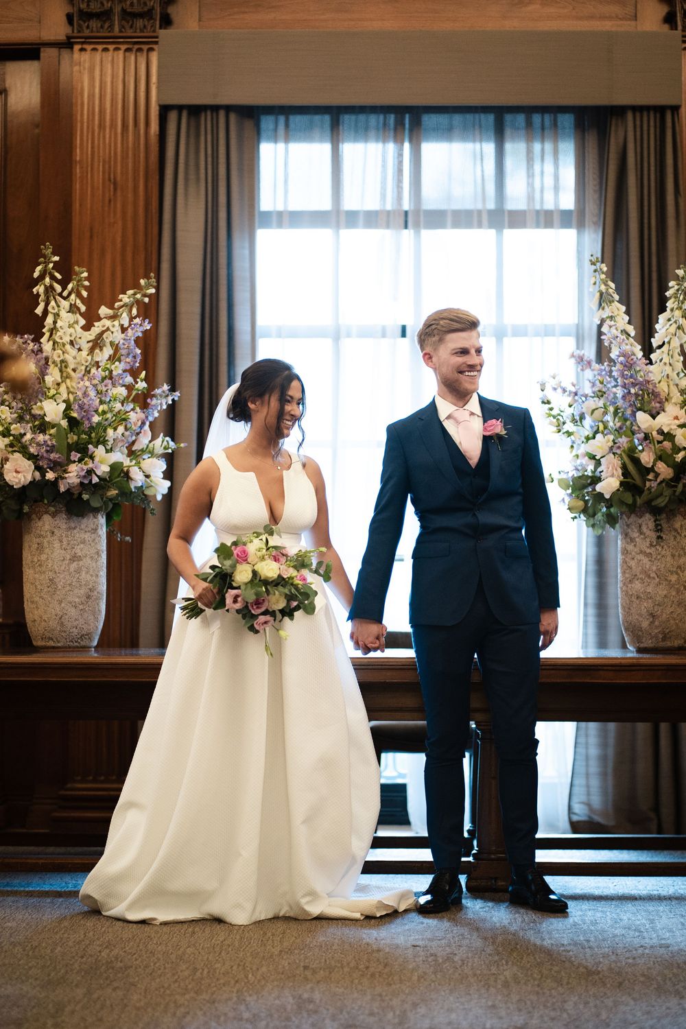 A bride and groom pose for photos in front of a window with beautiful floral arrangements.