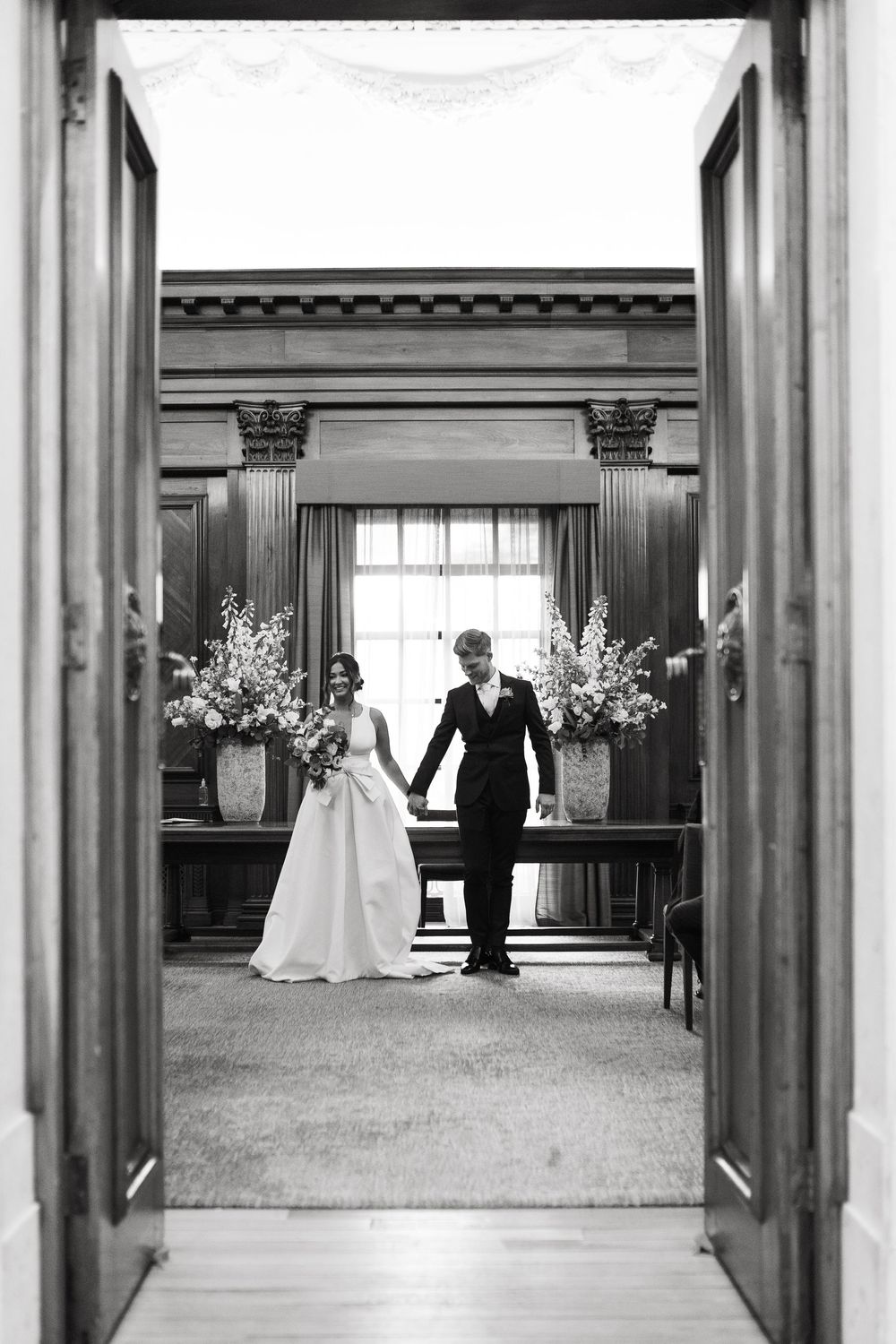 A black and white wedding photo capturing newlyweds holding hands in a doorway framed by floral arrangements.
