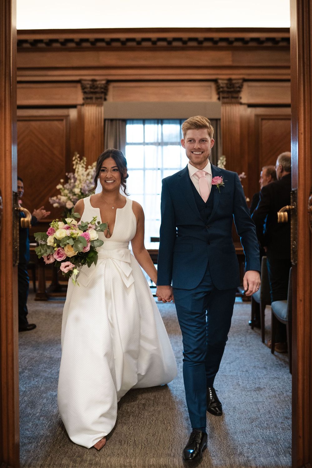 Newlyweds walk hand in hand through an elegant venue doorway with joyful expressions.