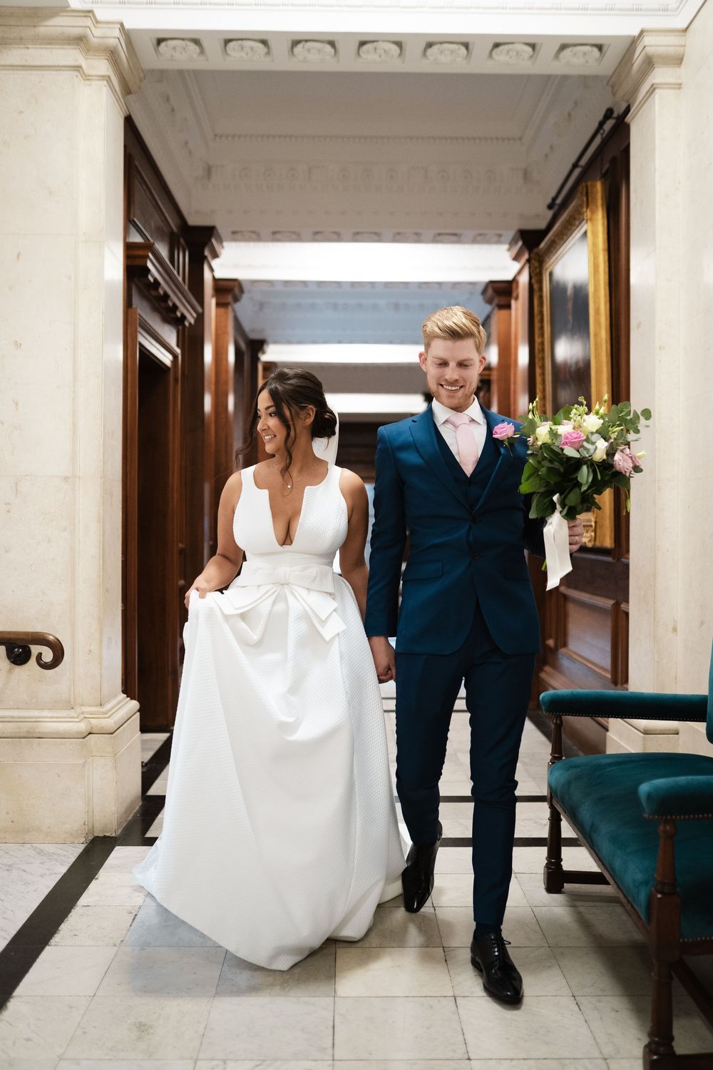 Couple in wedding attire walking through luxurious marble hallway with floral bouquet.