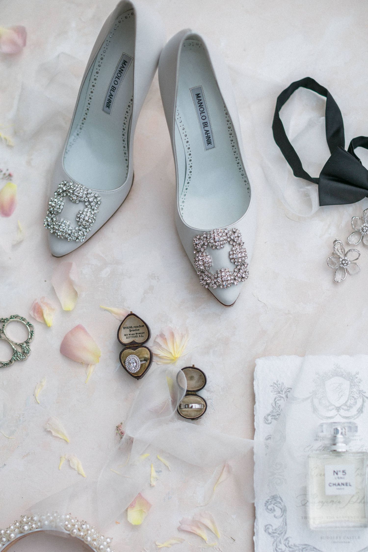 Elegant silver Manolo Blahnik shoes with crystal embellishments styled with Channel No 5 and rose petals on light marble surface flatlay
