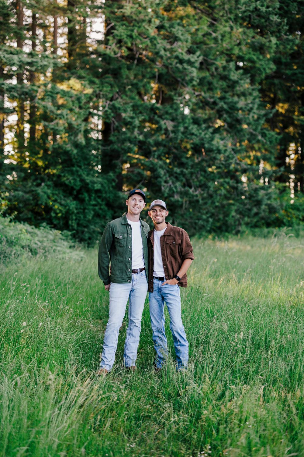 Two people stand together in a grassy meadow with tall evergreen trees in the background on a sunny day.
