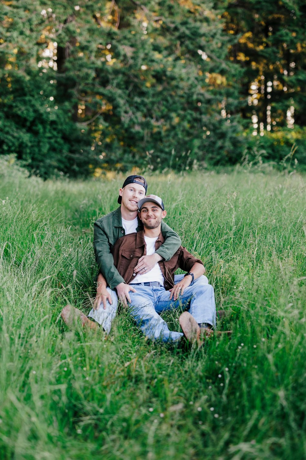 Two people sitting together in tall grass on a sunny day with green forest in the background.