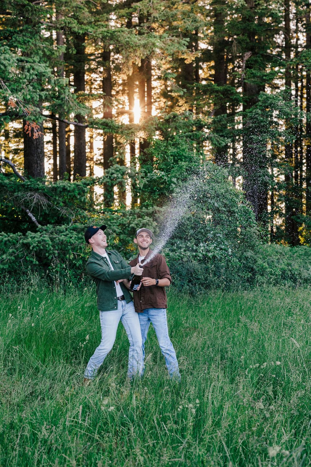 Two people spraying champagne in a sunlit forest clearing surrounded by tall evergreen trees in golden hour.