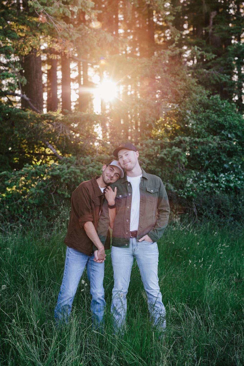 Two people in casual outfits embracing in a forest clearing with golden sunlight filtering through pine trees.