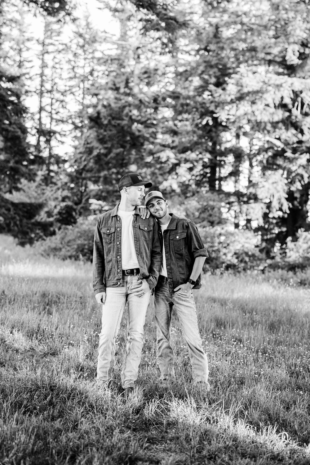 Two people in denim and jackets standing together in a grassy field with forest trees in the background.
