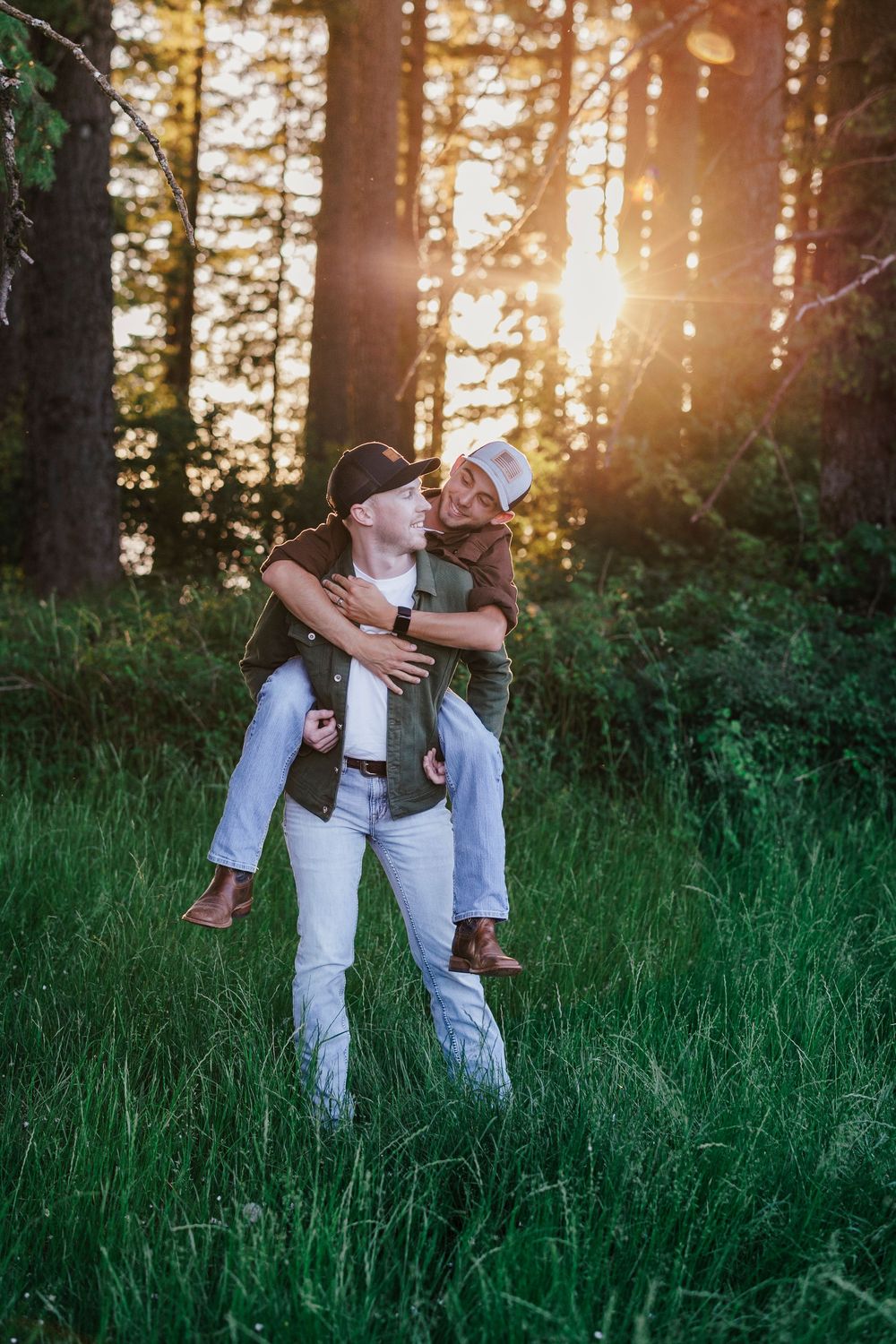 Two people embracing in tall grass during golden hour with sunlit pine trees in the background.