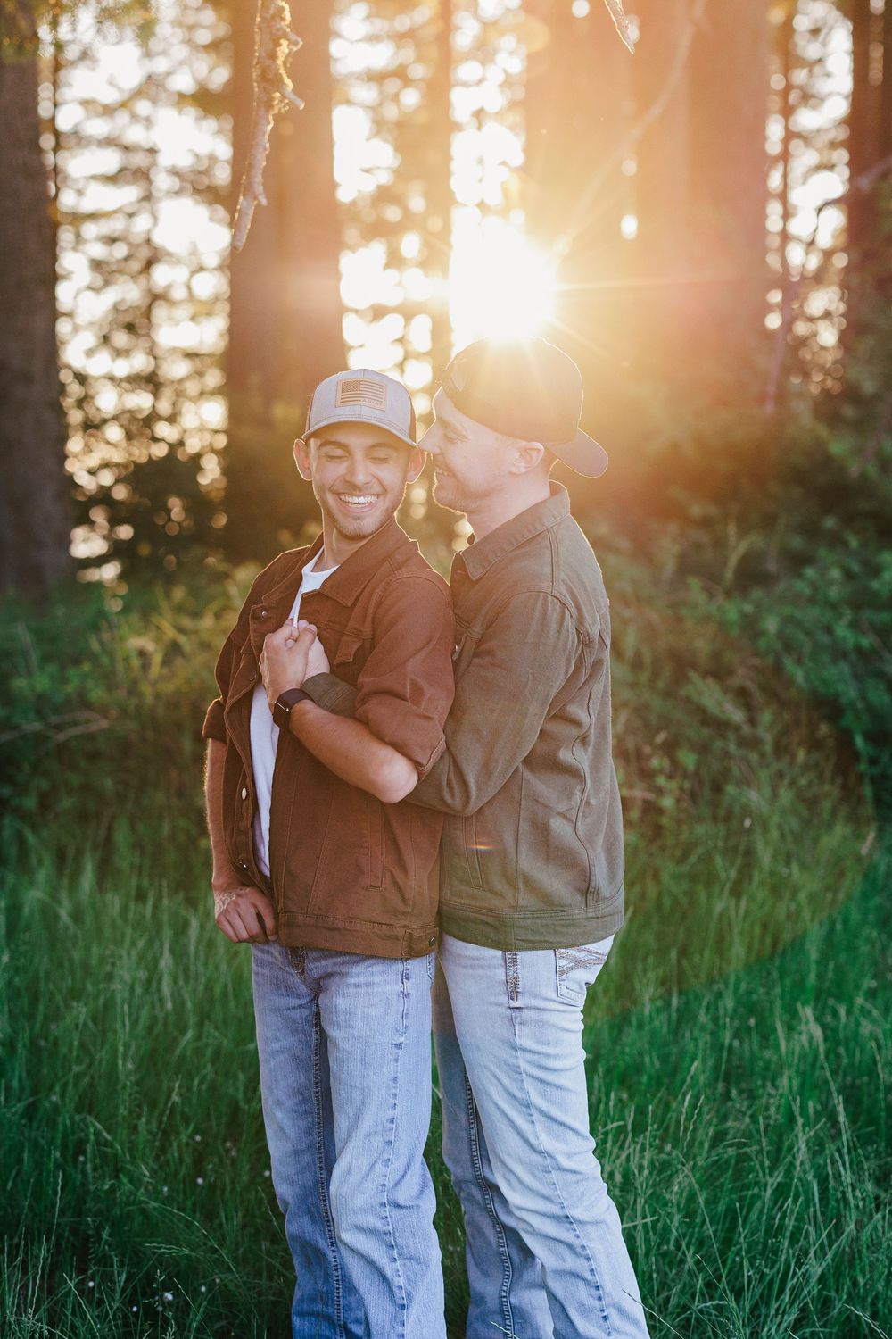 Two people share a warm embrace while standing in tall grass as golden sunlight streams through forest trees behind them.