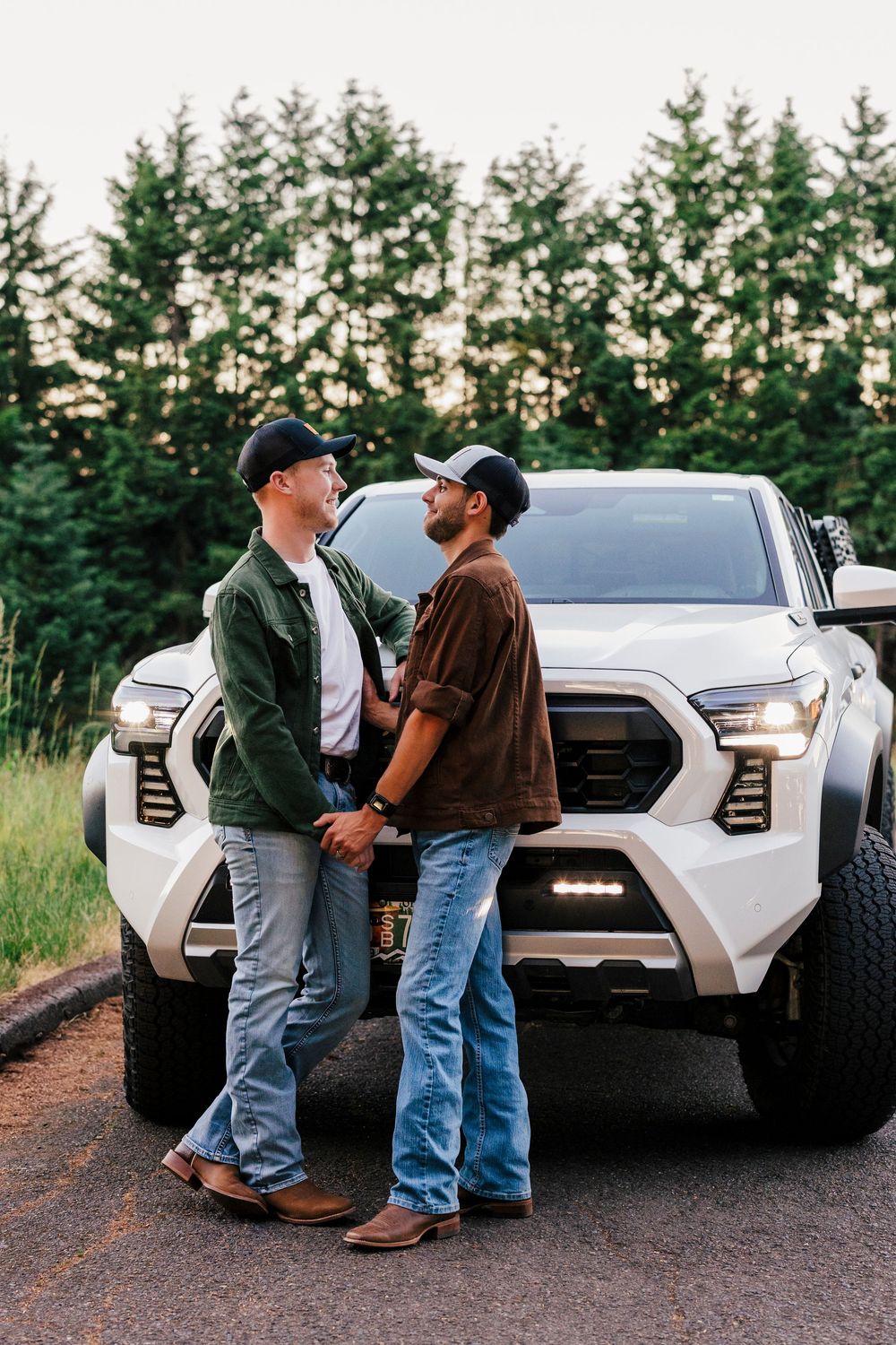 Two people in casual wear stand together in front of a custom white pickup truck with trees in the background at sunset.