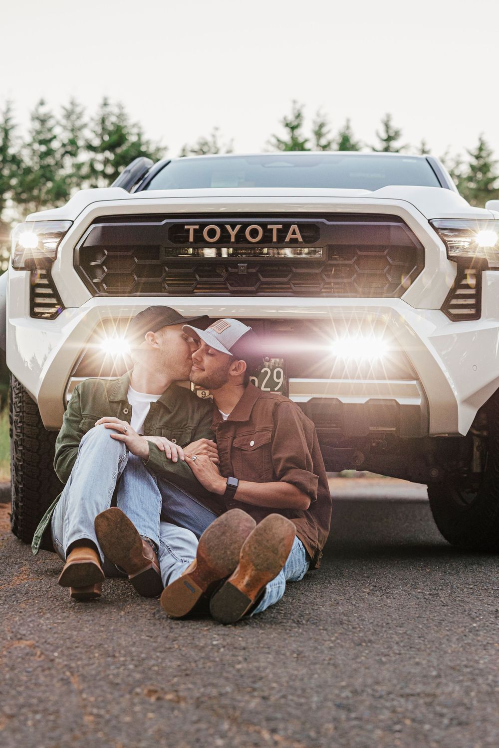 A couple sits in front of a white Toyota pickup truck with illuminated headlights at sunset.