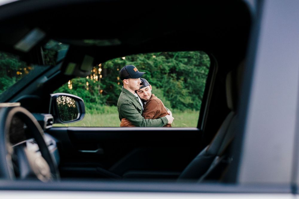 View through car window shows person in baseball cap drinking from a paper cup while standing outside.