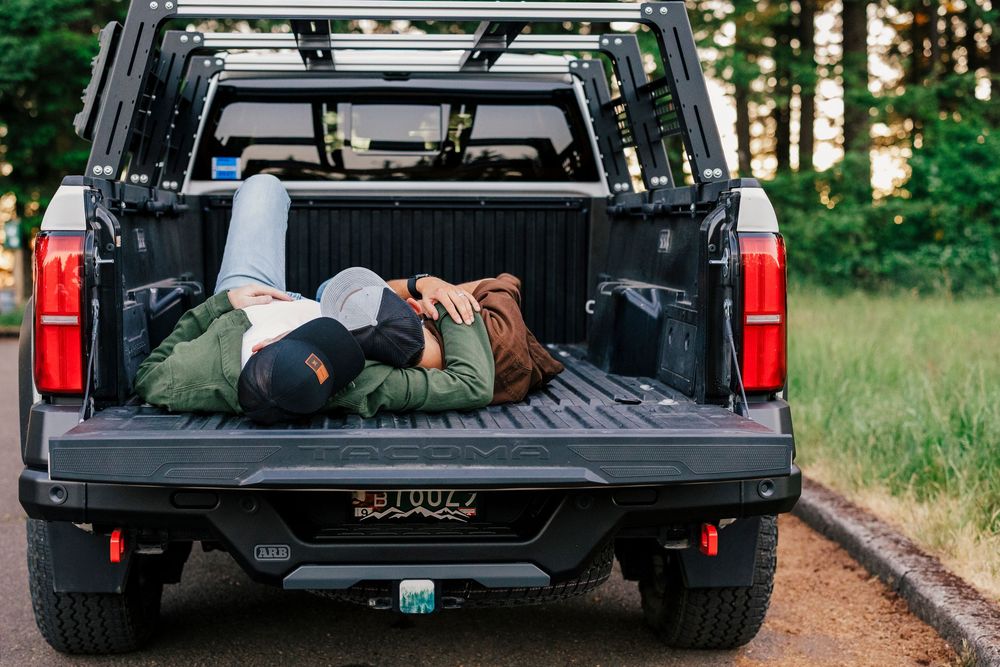Person resting in the bed of a black pickup truck parked on a forest road with camping gear and supplies.