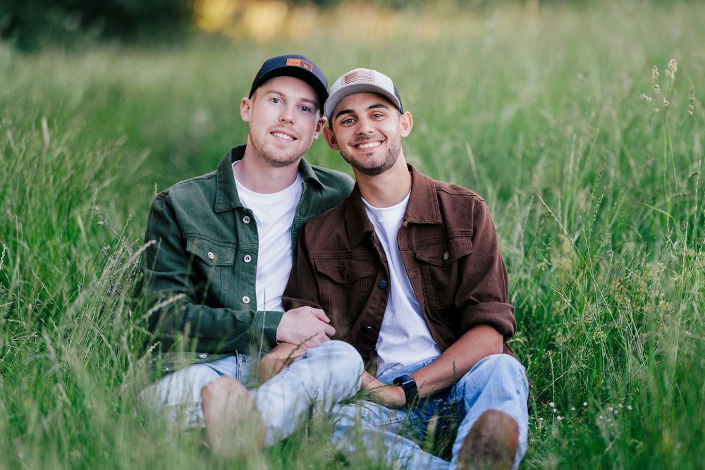 Two casually dressed people sitting together and smiling in tall grass on a sunny day.