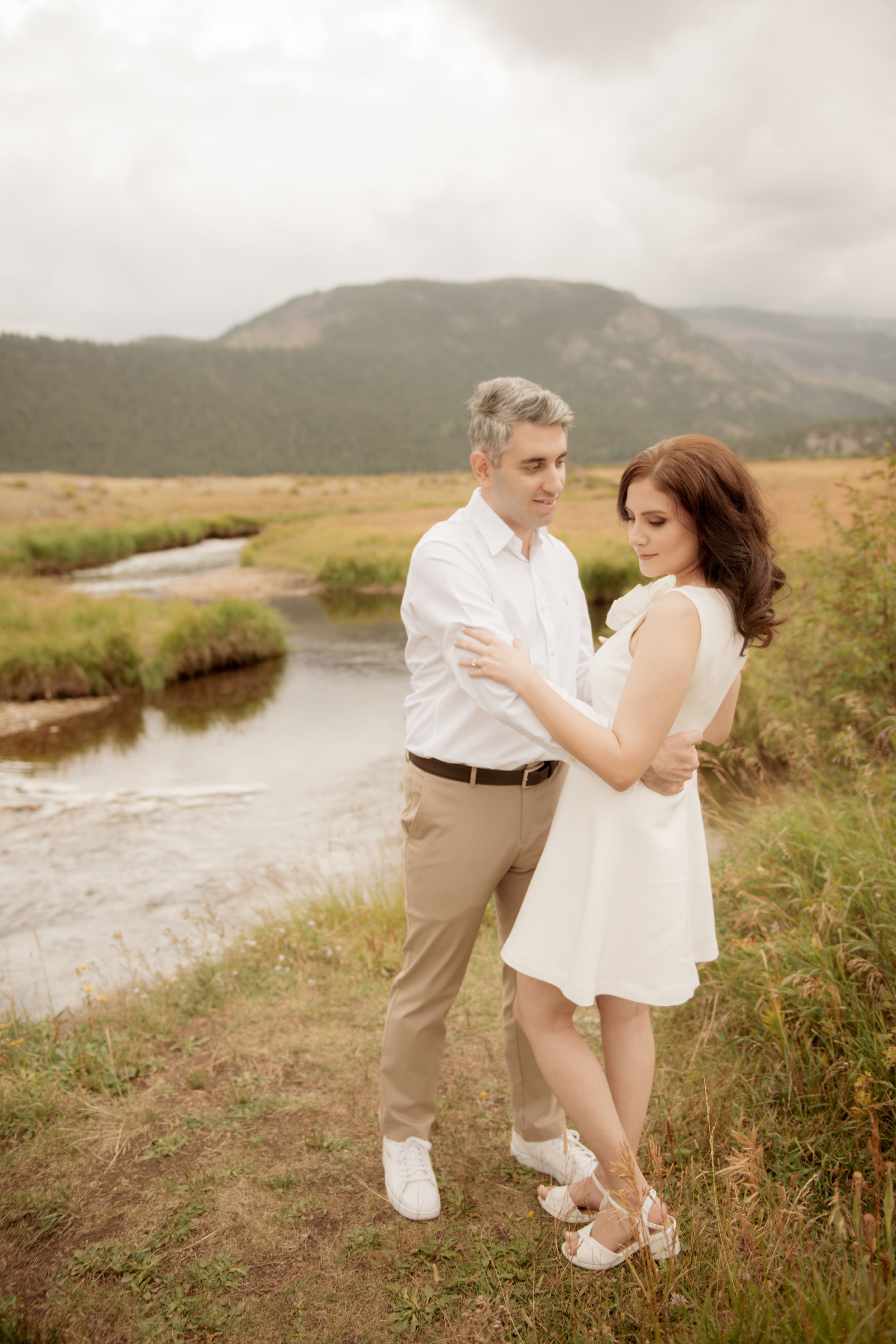 A couple embraces on a hillside near a winding stream with mountains in the background during golden hour.