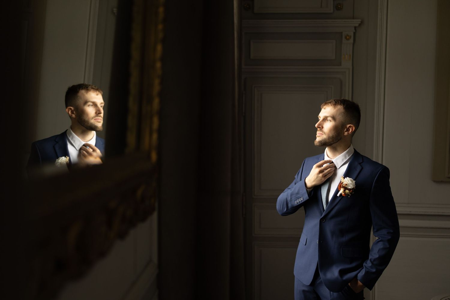 Man in navy suit adjusts tie while looking in mirror with dramatic side lighting.