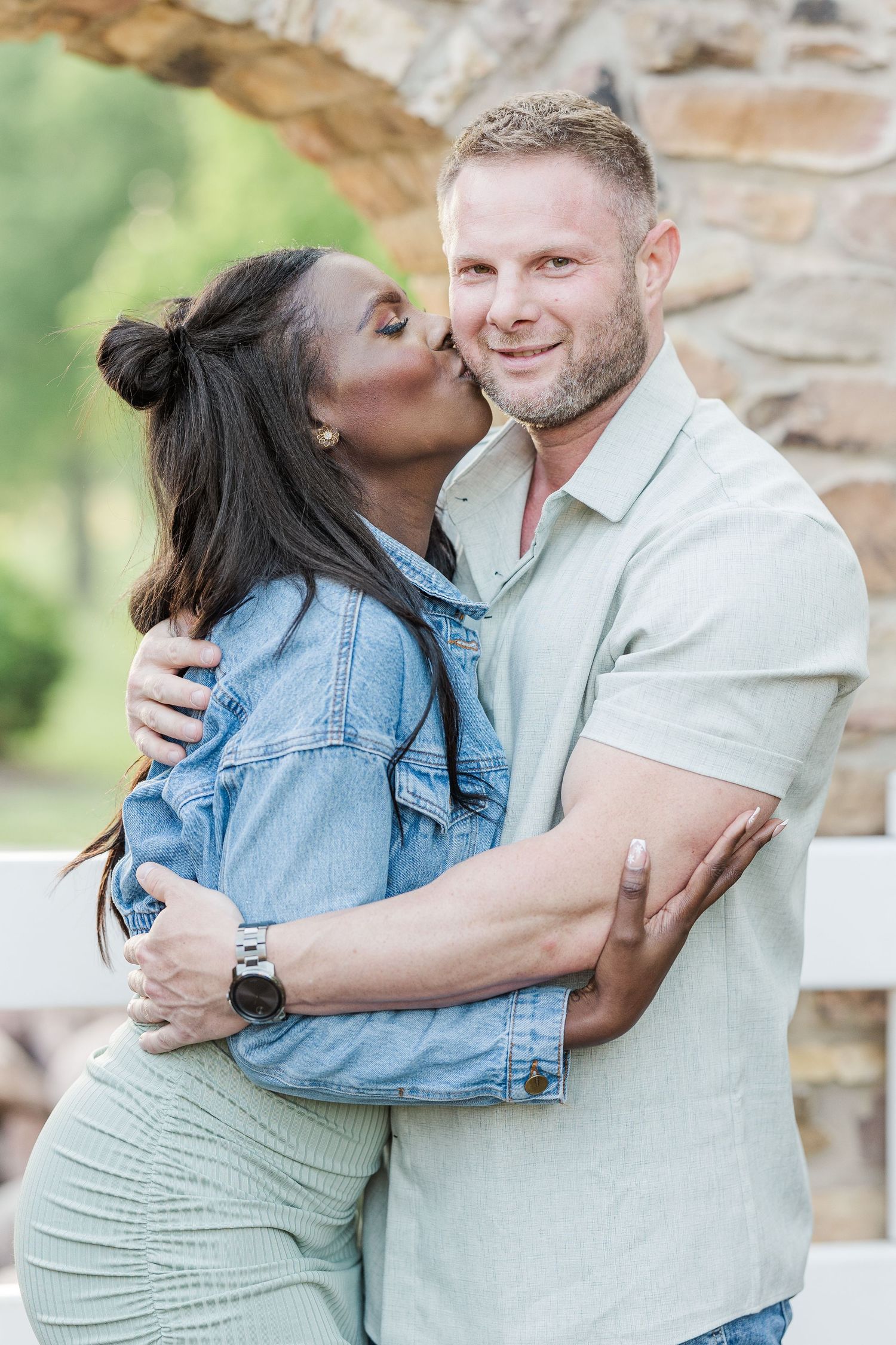A romantic outdoor photo showing a couple embracing, with a woman in denim jacket kissing a man in light colored shirt.