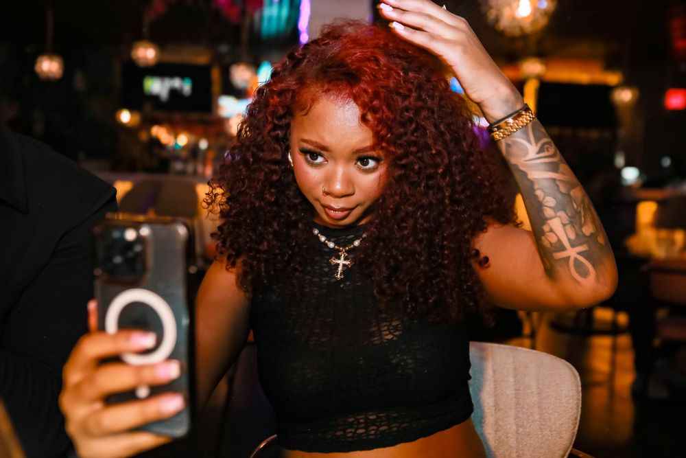 A person with curly red hair smiles while taking a selfie in a dimly lit venue.