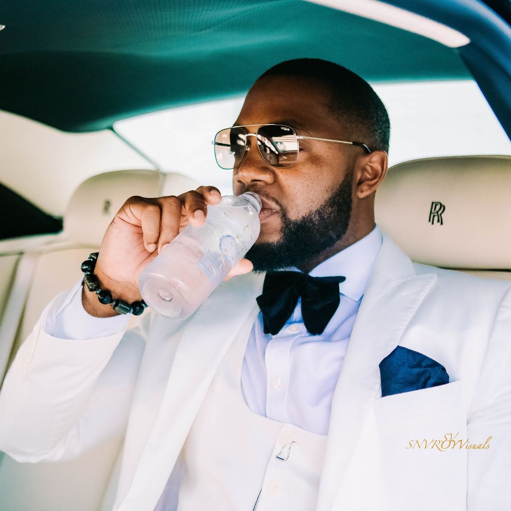 Person in white tuxedo and bowtie drinking water in luxury car interior.