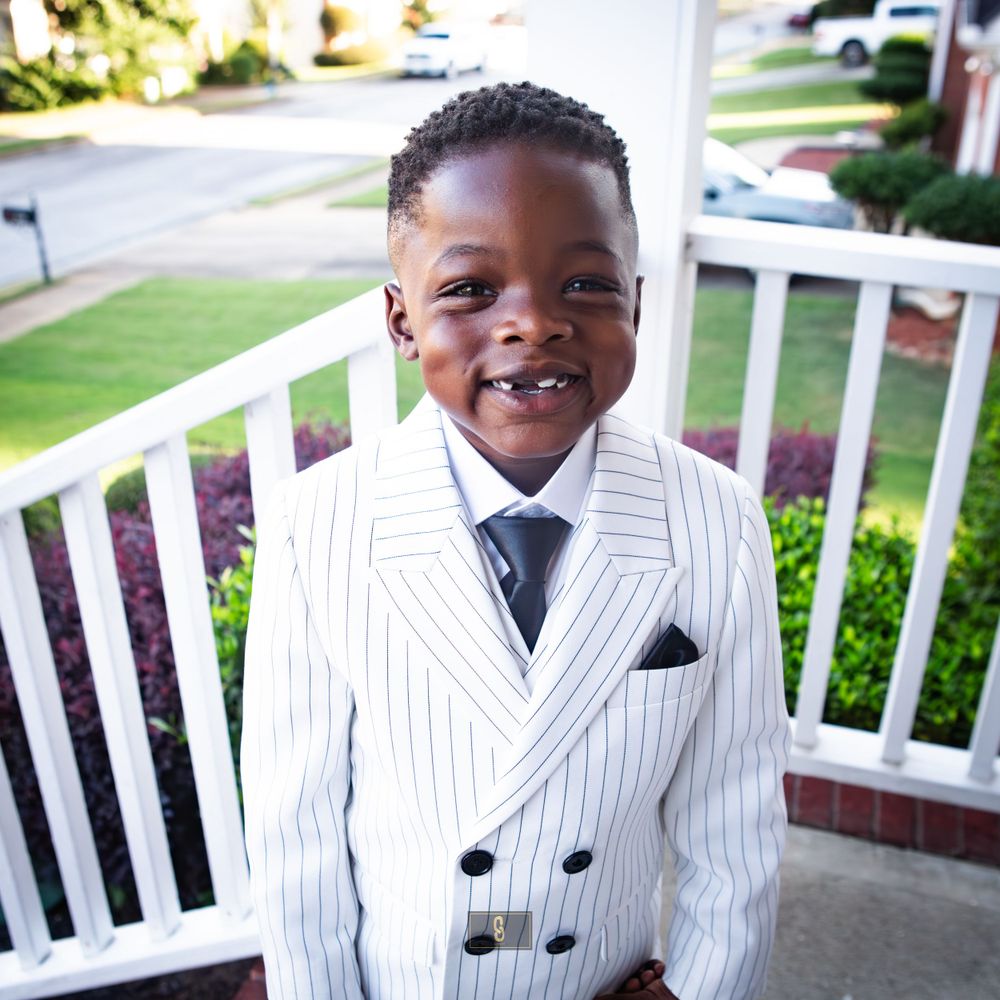 A young person in a white pinstripe suit smiles brightly on an outdoor porch.