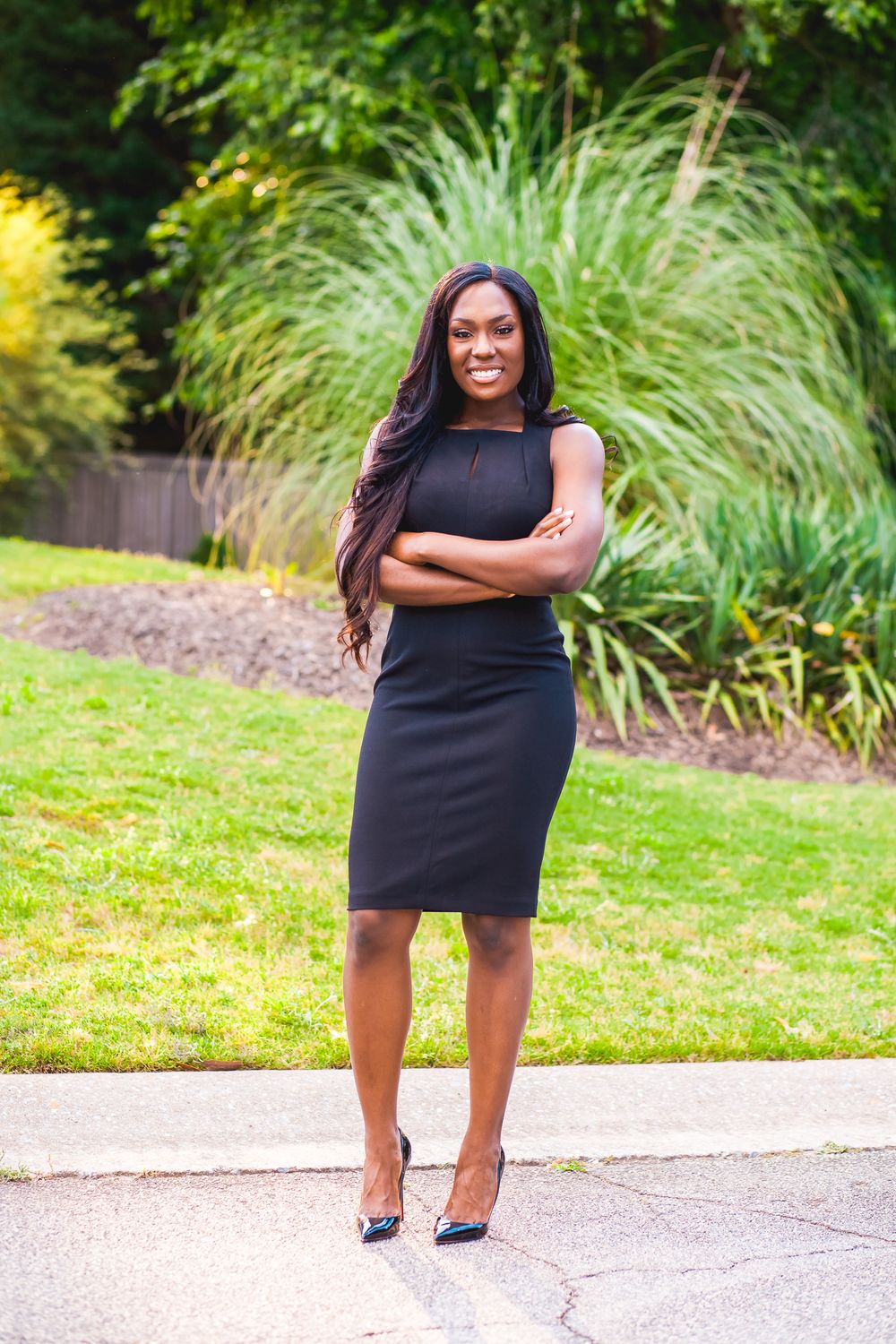 Full length portrait in a fitted black dress and heels with landscaped garden background.