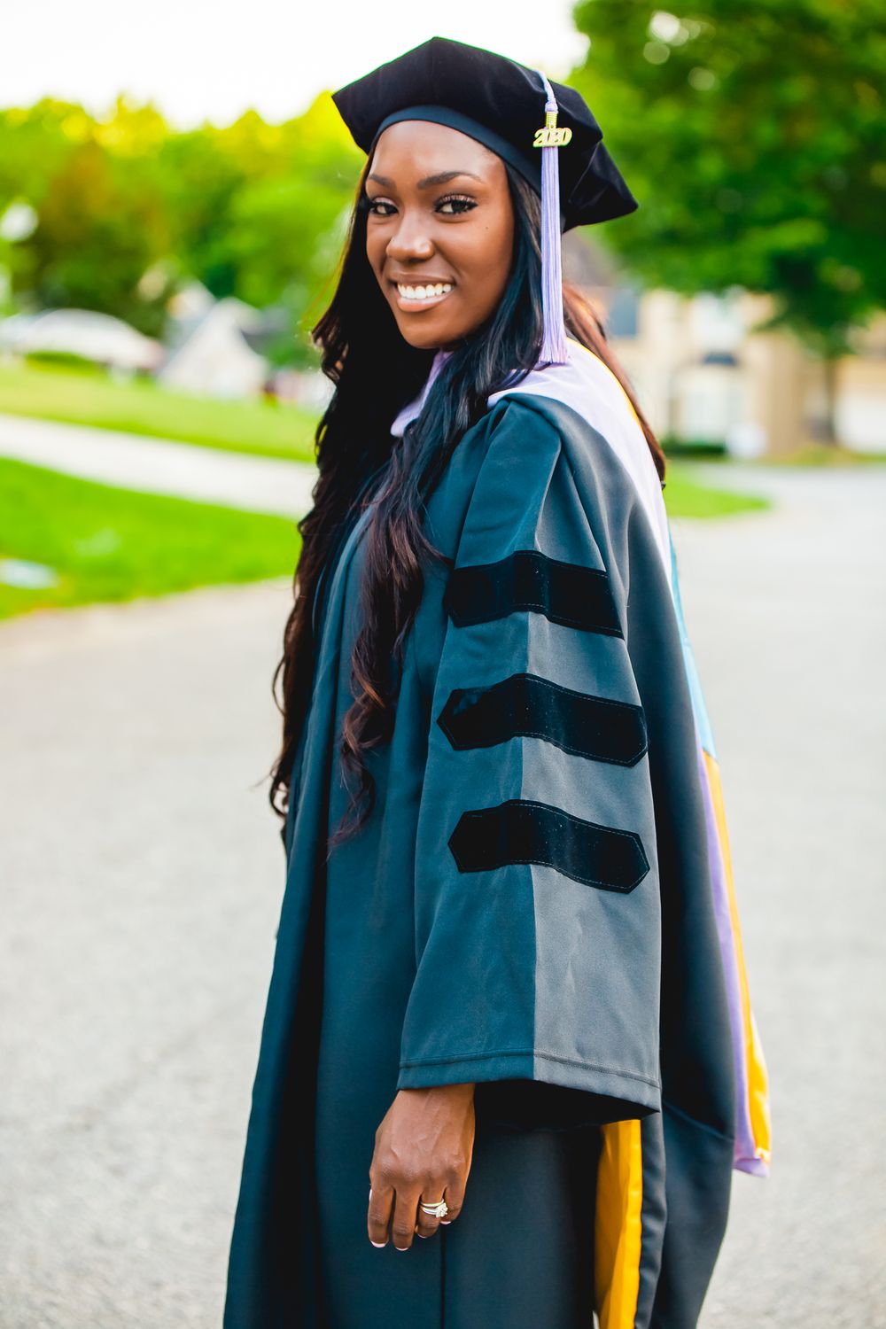 Graduate wearing traditional doctoral robe with velvet stripes poses outdoors on graduation day.