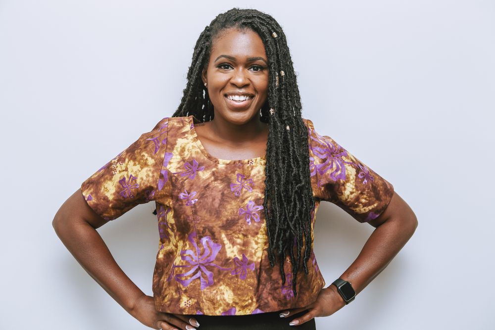 Someone wearing a floral patterned top with long braided hair poses with hands on hips against white background.