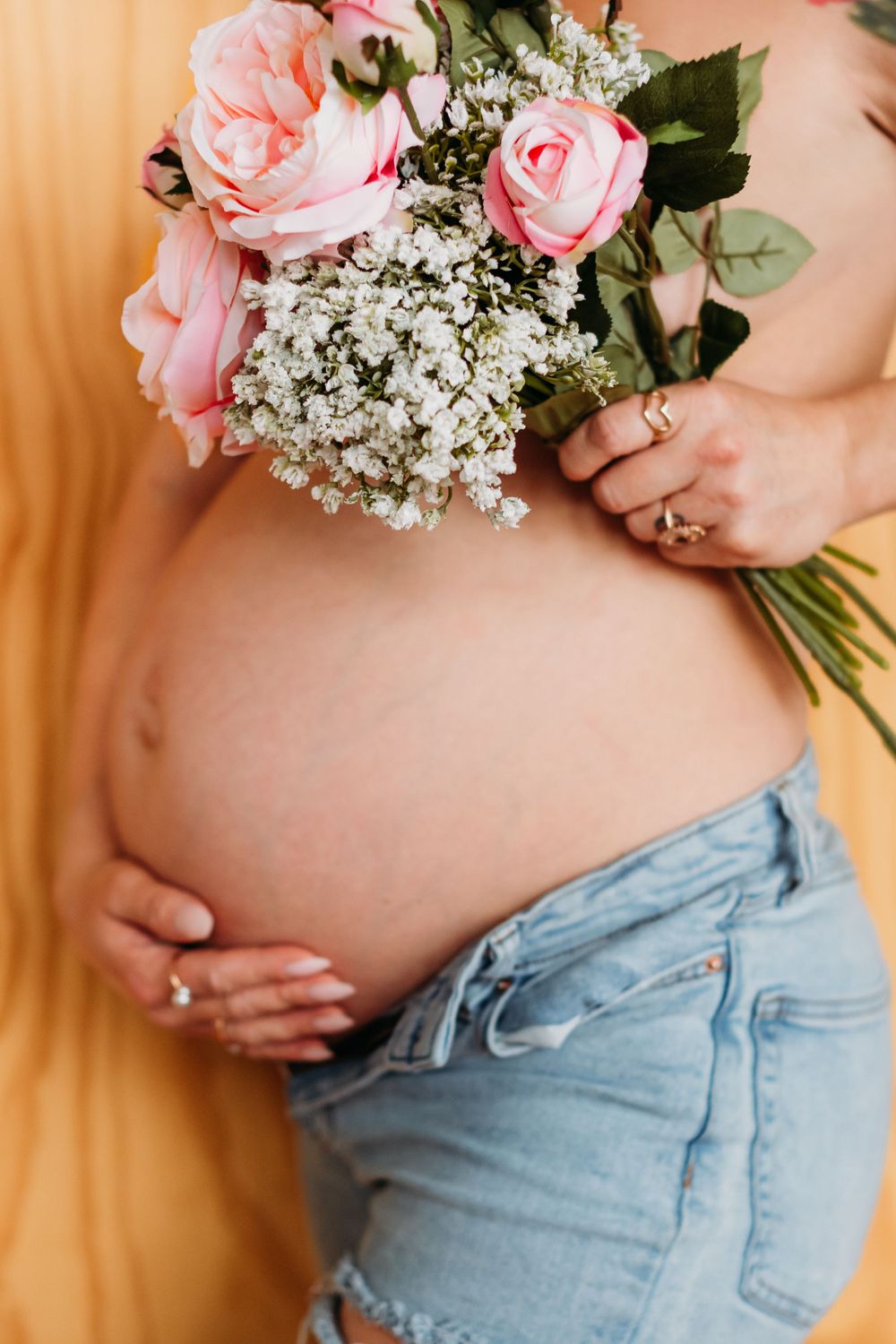 Pregnant belly with pink roses and baby's breath bouquet held against light denim jeans and peach background.