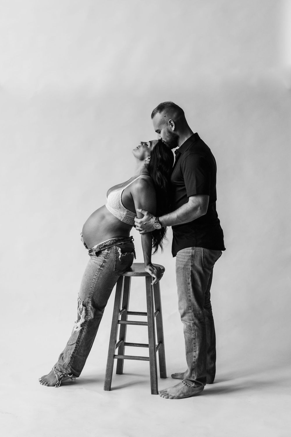 A black and white artistic maternity photo featuring a couple posing intimately with a wooden stool in a studio setting.