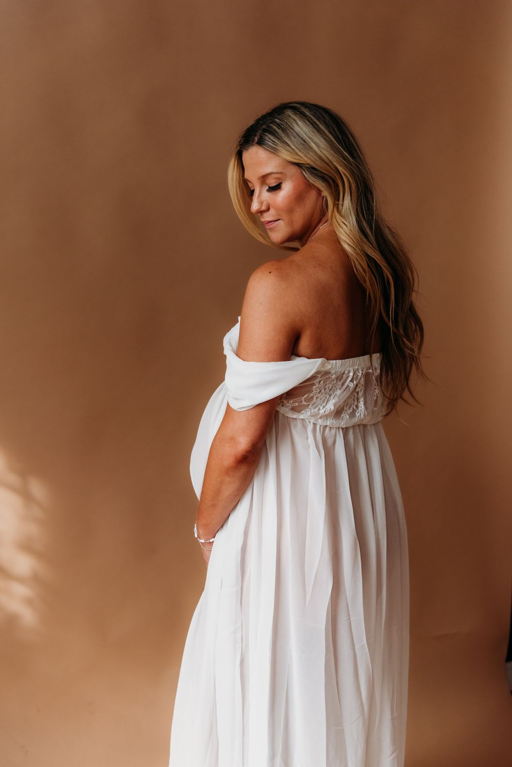 Side view portrait in white off-shoulder flowing dress against warm brown background with soft shadows.