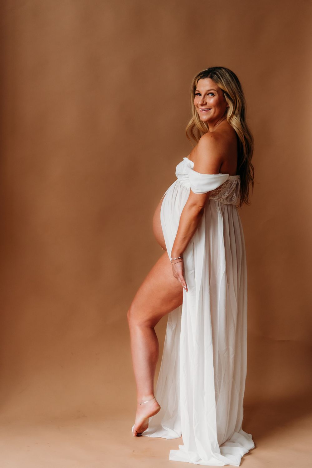 A pregnant woman in a flowing white off-shoulder dress poses elegantly against a brown backdrop during a maternity photoshoot.