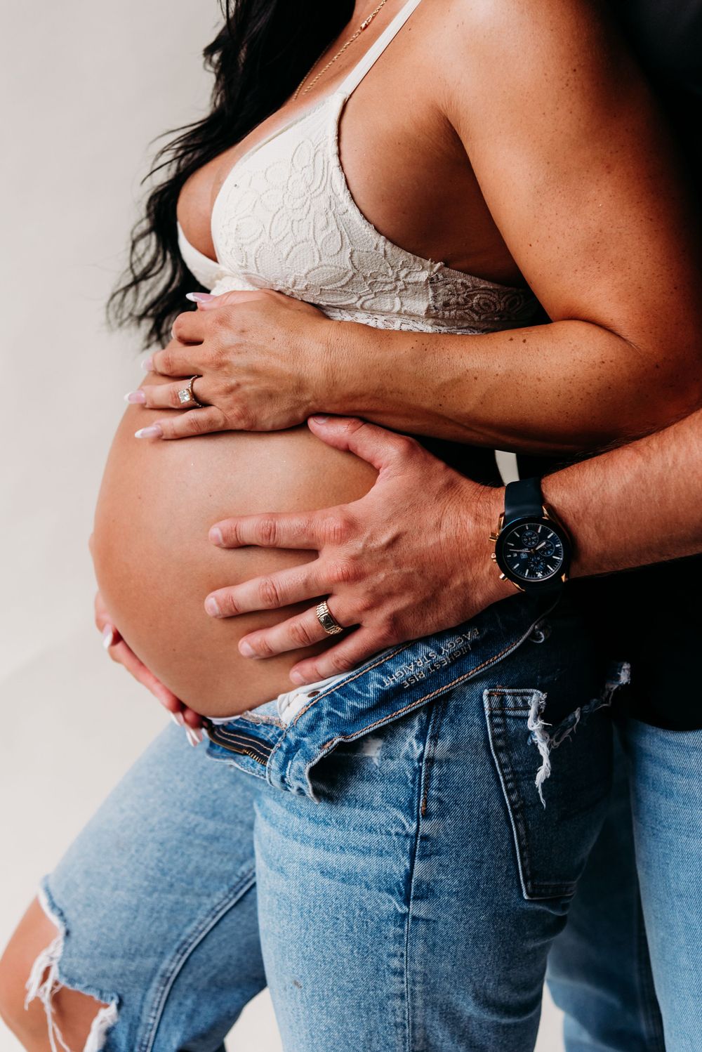 Close up of a pregnant belly being held by hands, one wearing a black watch and ring, against distressed jeans.