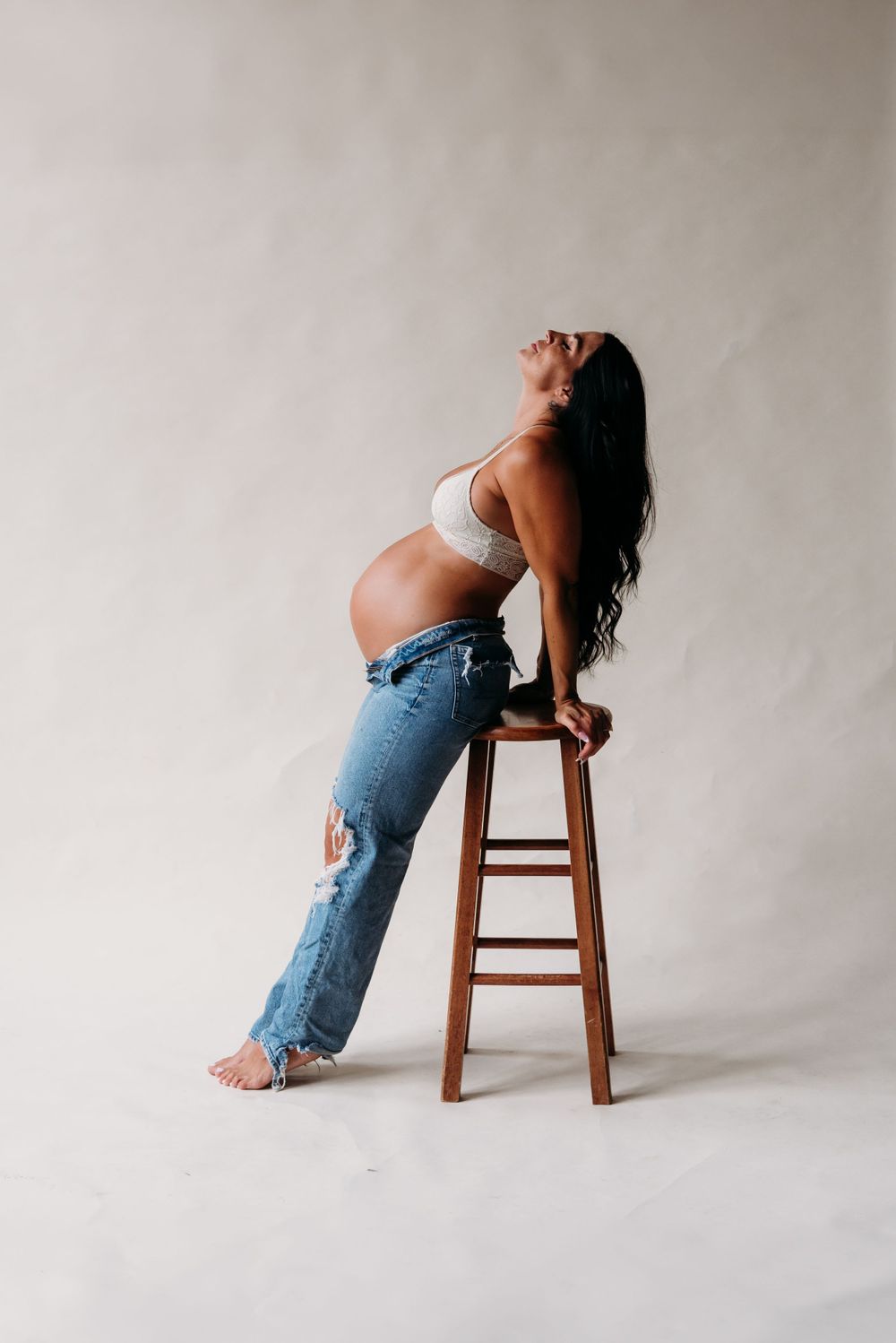 A pregnant woman leans back on a wooden stool wearing distressed jeans and a white top against a light backdrop.