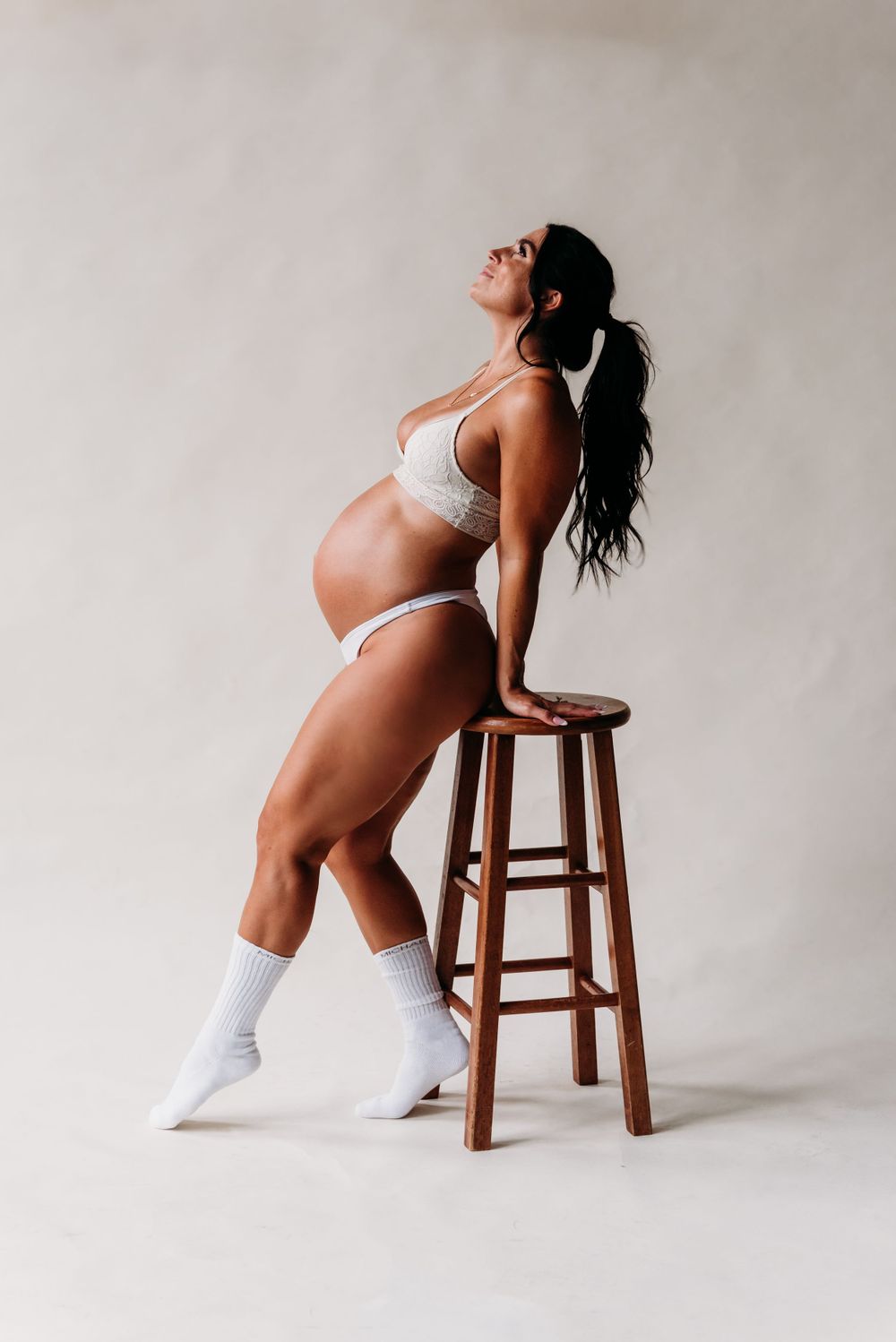 Maternity photo in white lingerie showing woman leaning against wooden stool in artistic pose against neutral backdrop.