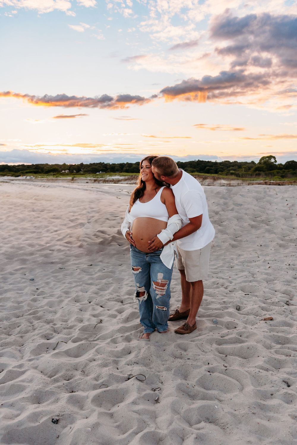 A couple embraces on a sandy beach at sunset with beautiful orange sky and clouds in the background.