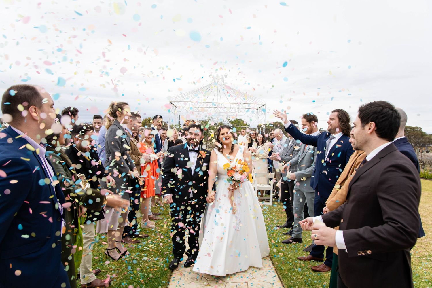 Wedding guests celebrate with confetti as newlyweds exit their outdoor ceremony.