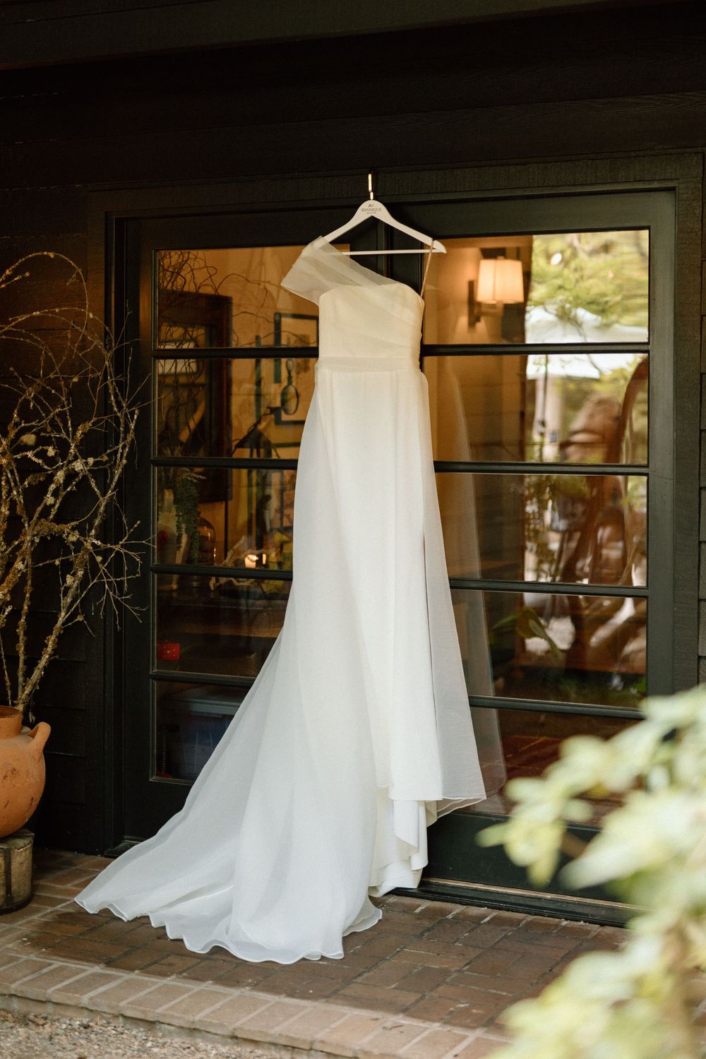 An elegant white wedding dress hangs on display against a dark green window frame with natural light filtering through.