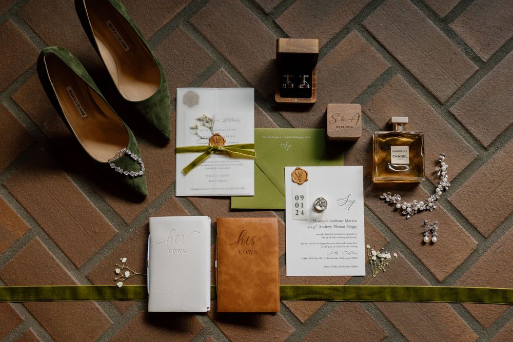 Wedding invitation suite with green ribbon, perfume bottle, jewelry and shoes arranged on brown tile flooring.