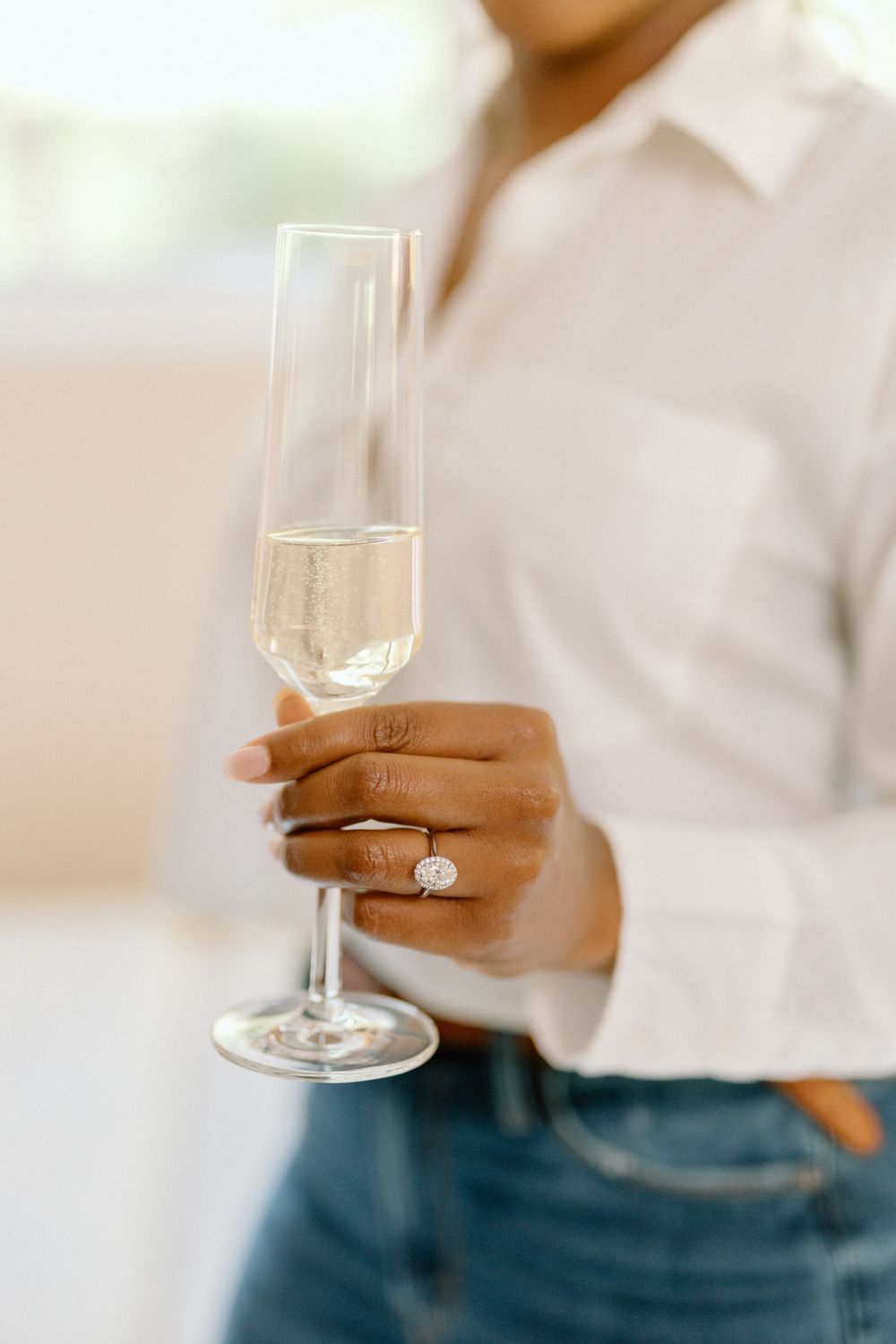 Hand holding a champagne flute with a sparkling diamond engagement ring visible on the finger.