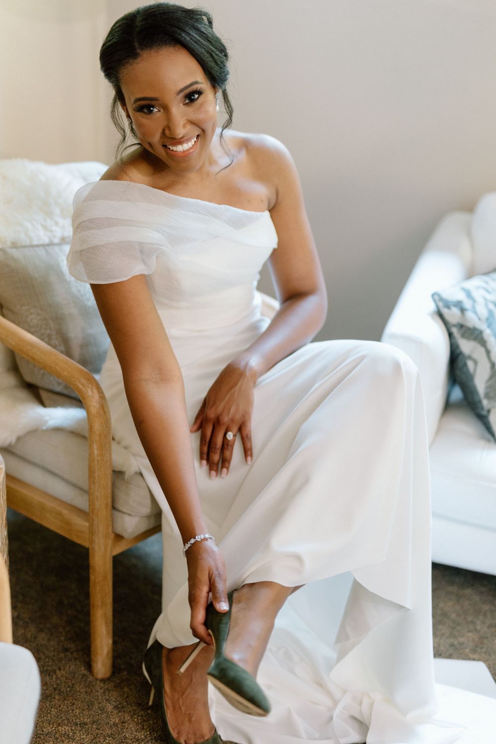 A bride in an elegant off-shoulder white gown adjusts her shoe while sitting in a cozy interior setting.