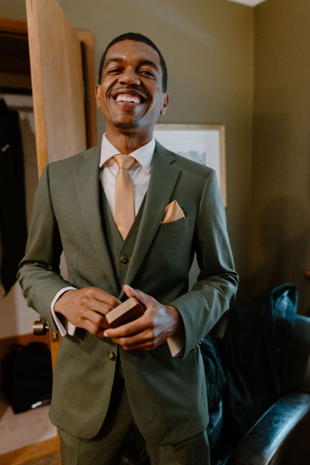 A person wearing a sage green three-piece suit with a peach tie and pocket square smiles warmly while getting dressed.