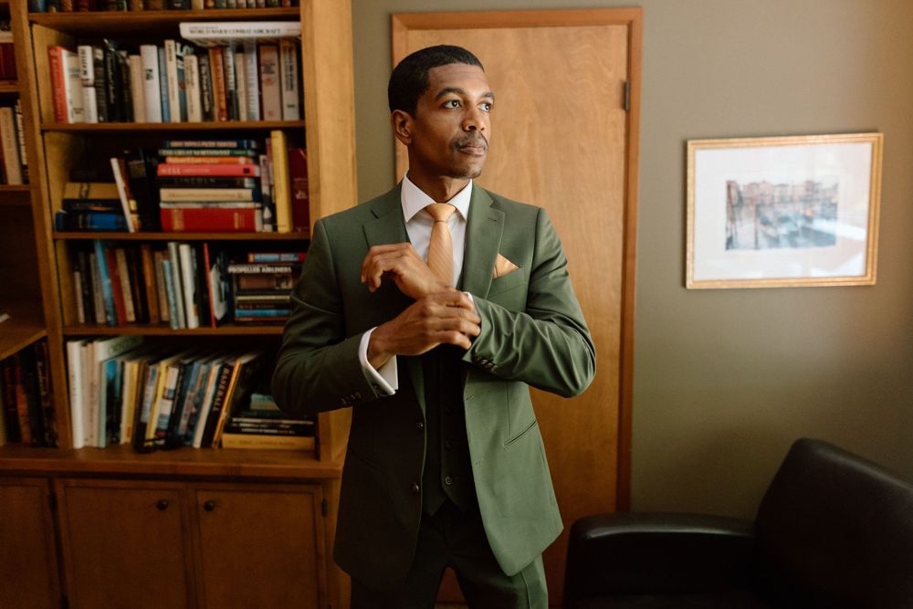 Professional wearing a green blazer and black tie adjusts his collar while standing in an office with bookshelves.