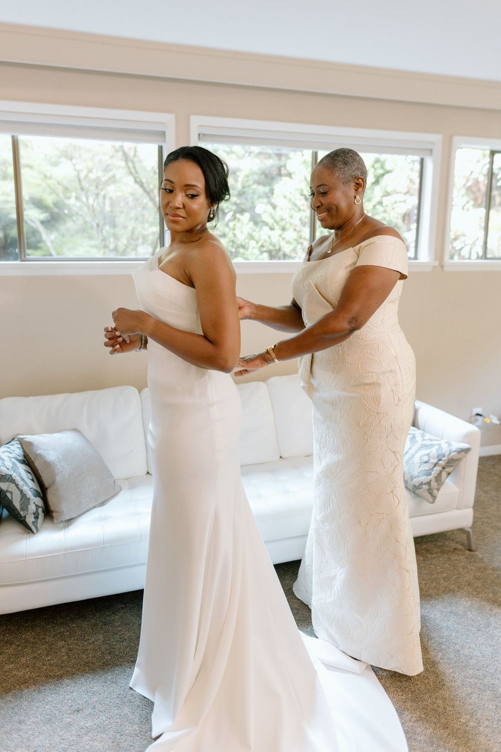 A helper assists with zipping up an elegant white wedding dress in a sunlit room with a white couch.