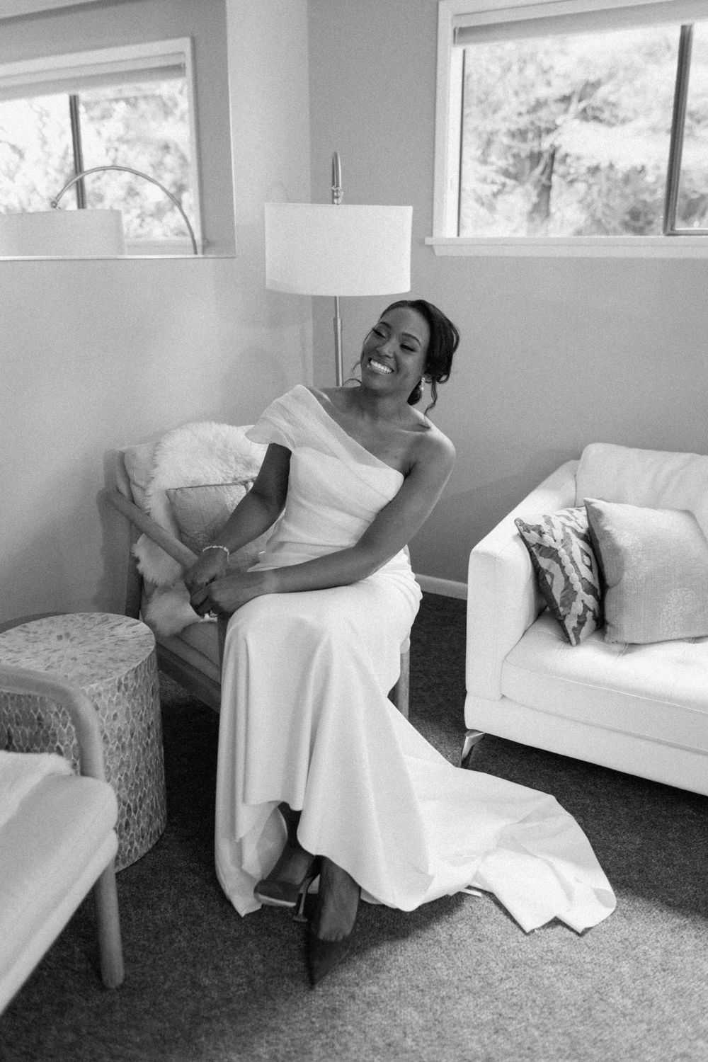 A stunning wedding scene shows a bride in an elegant white off-shoulder gown relaxing in a bright, airy room.