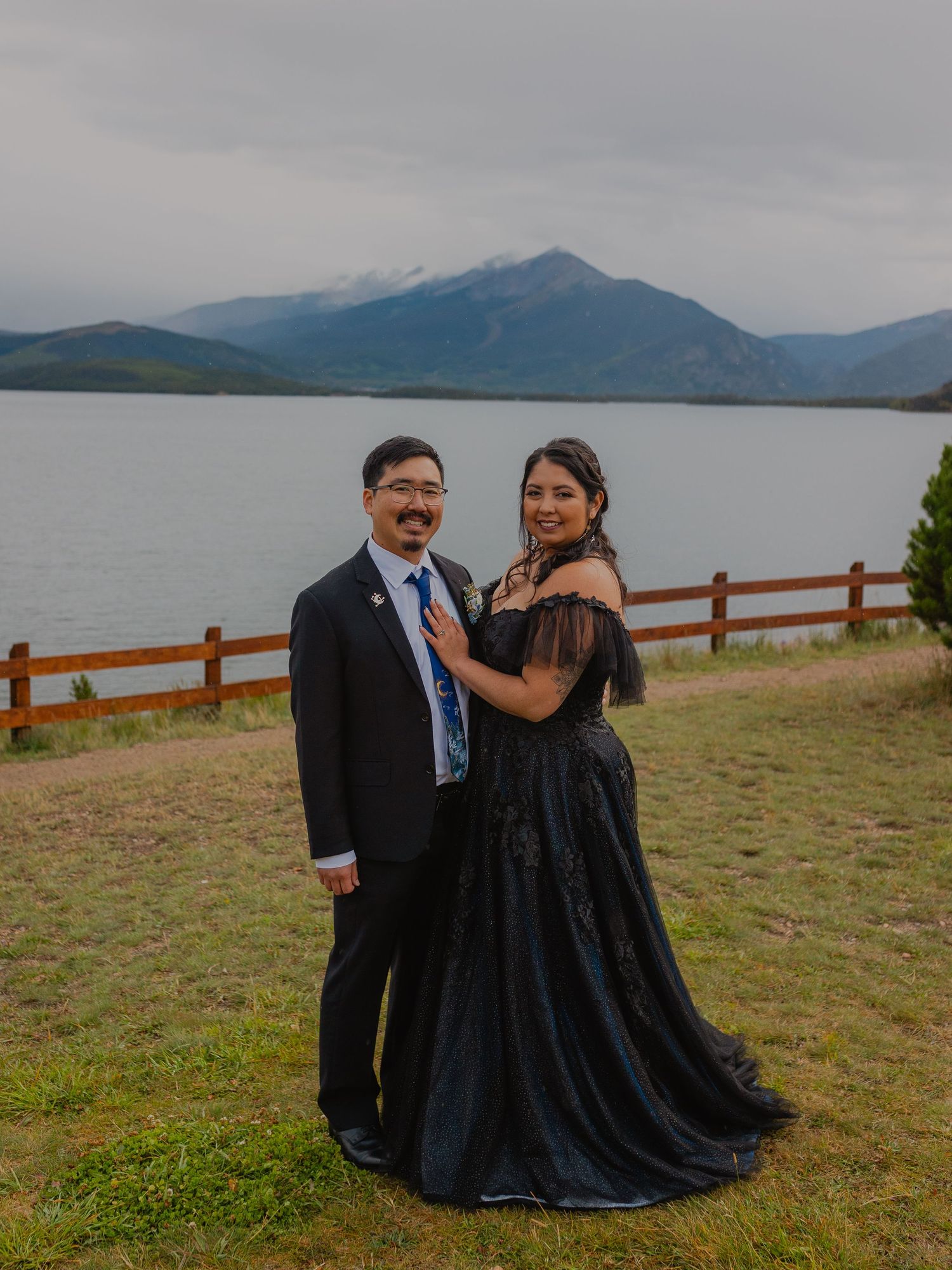 Elegant couple in formal attire poses near lake with mountain backdrop and wooden fence.