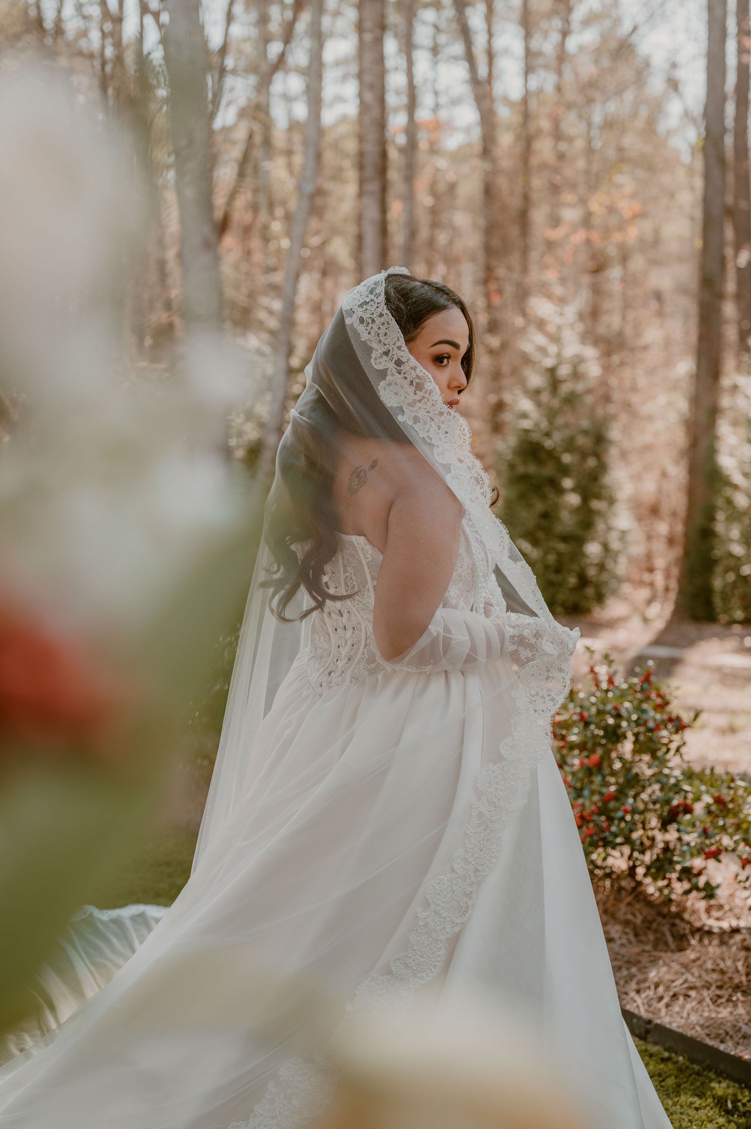 A bride in a flowing lace veil and white wedding gown poses in an autumn forest setting during golden hour.