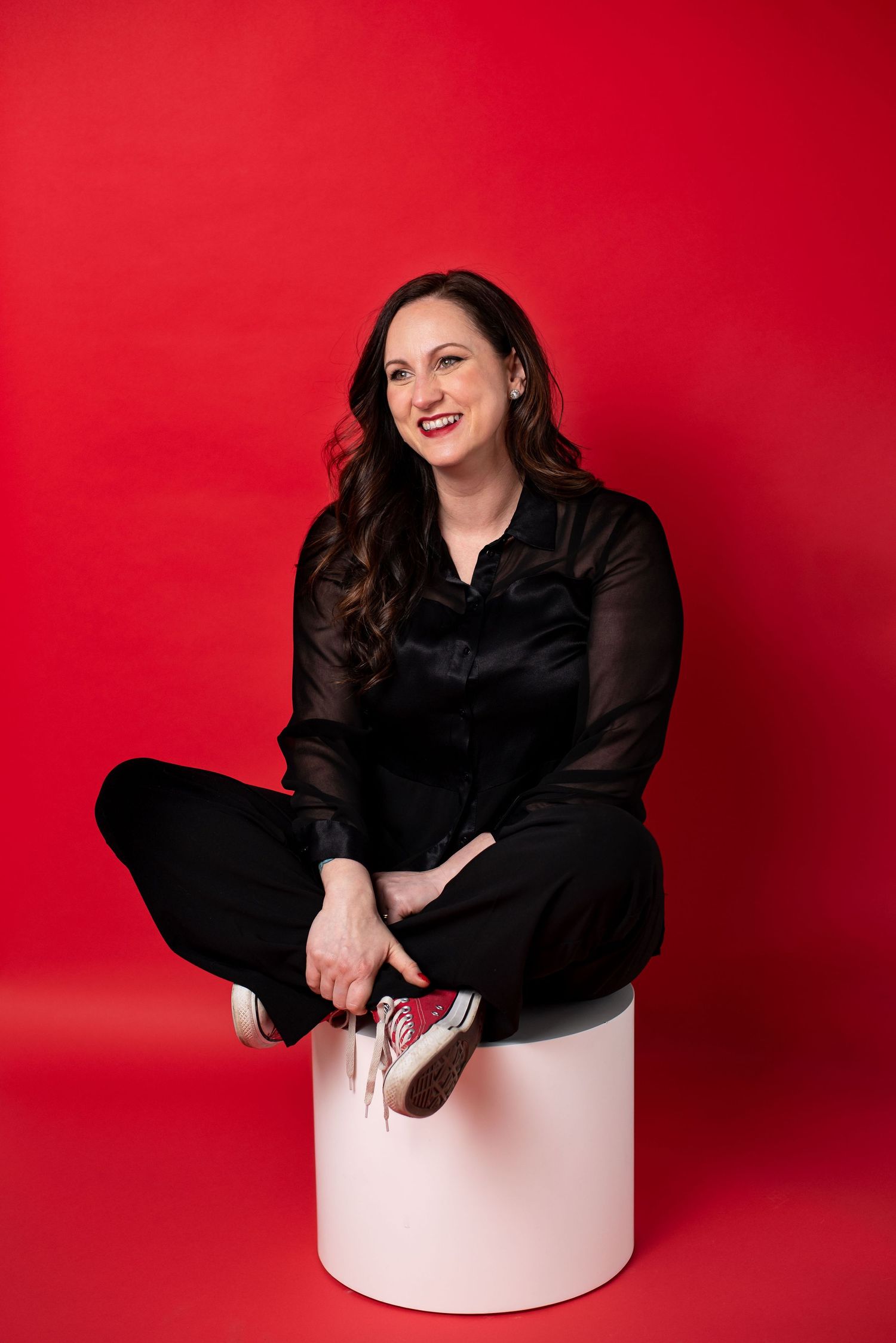 A person in a black outfit sits cross-legged on a pink cylindrical stool against a vibrant red backdrop.