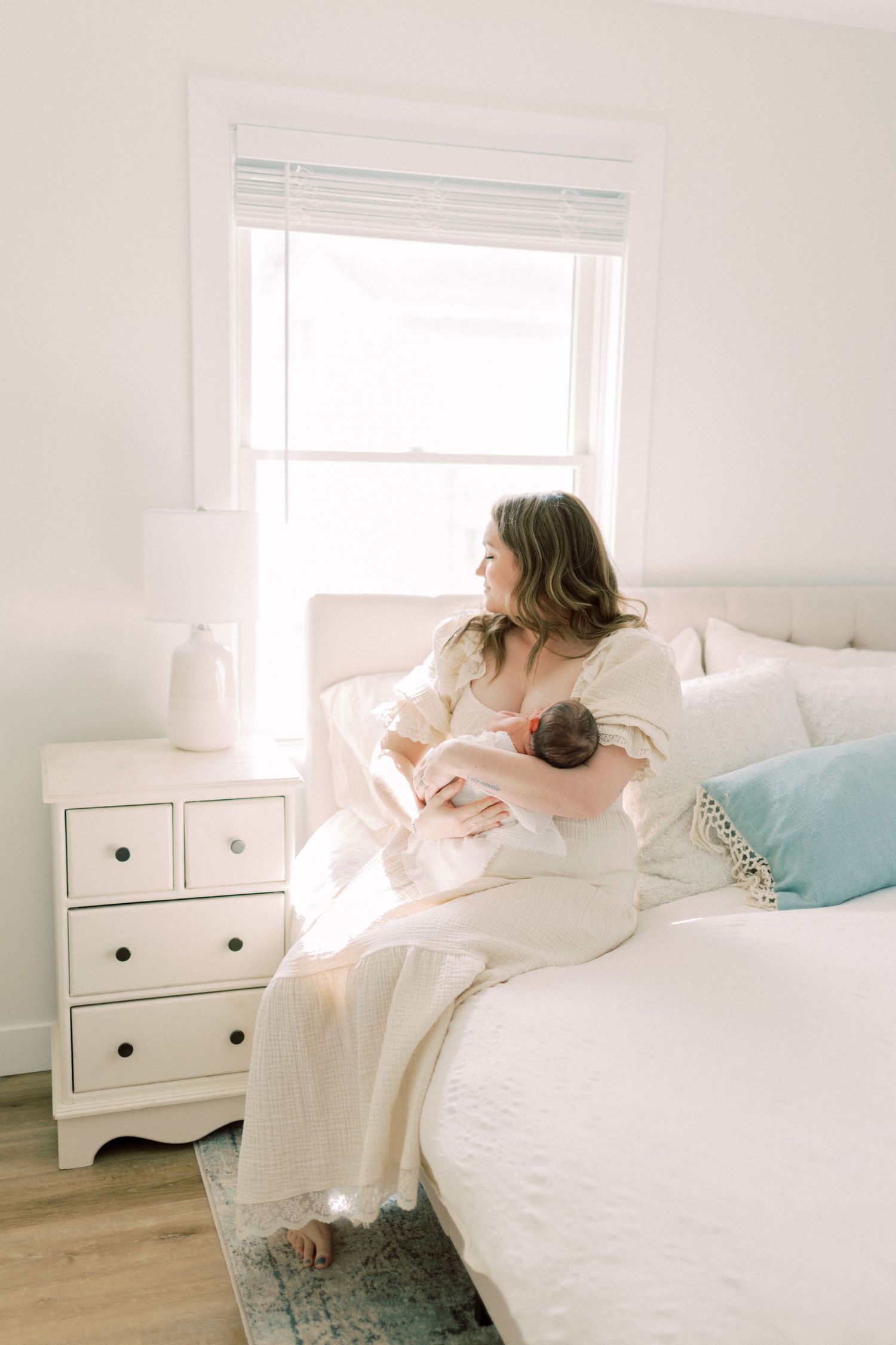 Soft morning light streams through window onto white bed where mother cradles baby in peaceful bedroom setting.